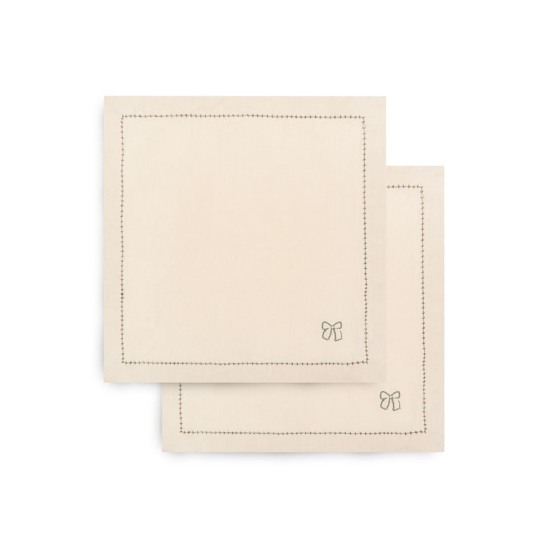 Two square cream-colored cotton napkins with stitched borders and a small bow motif in the lower right corner, stacked on a white background.