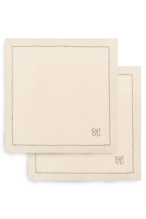 Two square cream-colored cotton napkins with stitched borders and a small bow motif in the lower right corner, stacked on a white background.