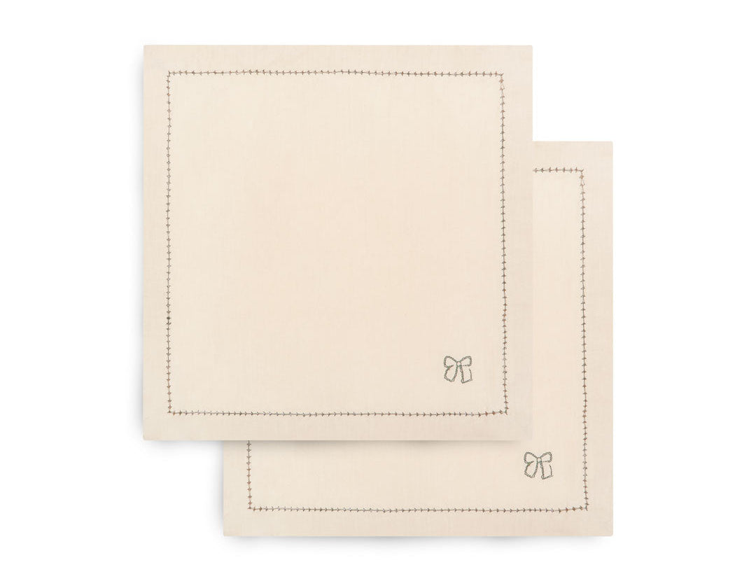 Two square cream-colored cotton napkins with stitched borders and a small bow motif in the lower right corner, stacked on a white background.