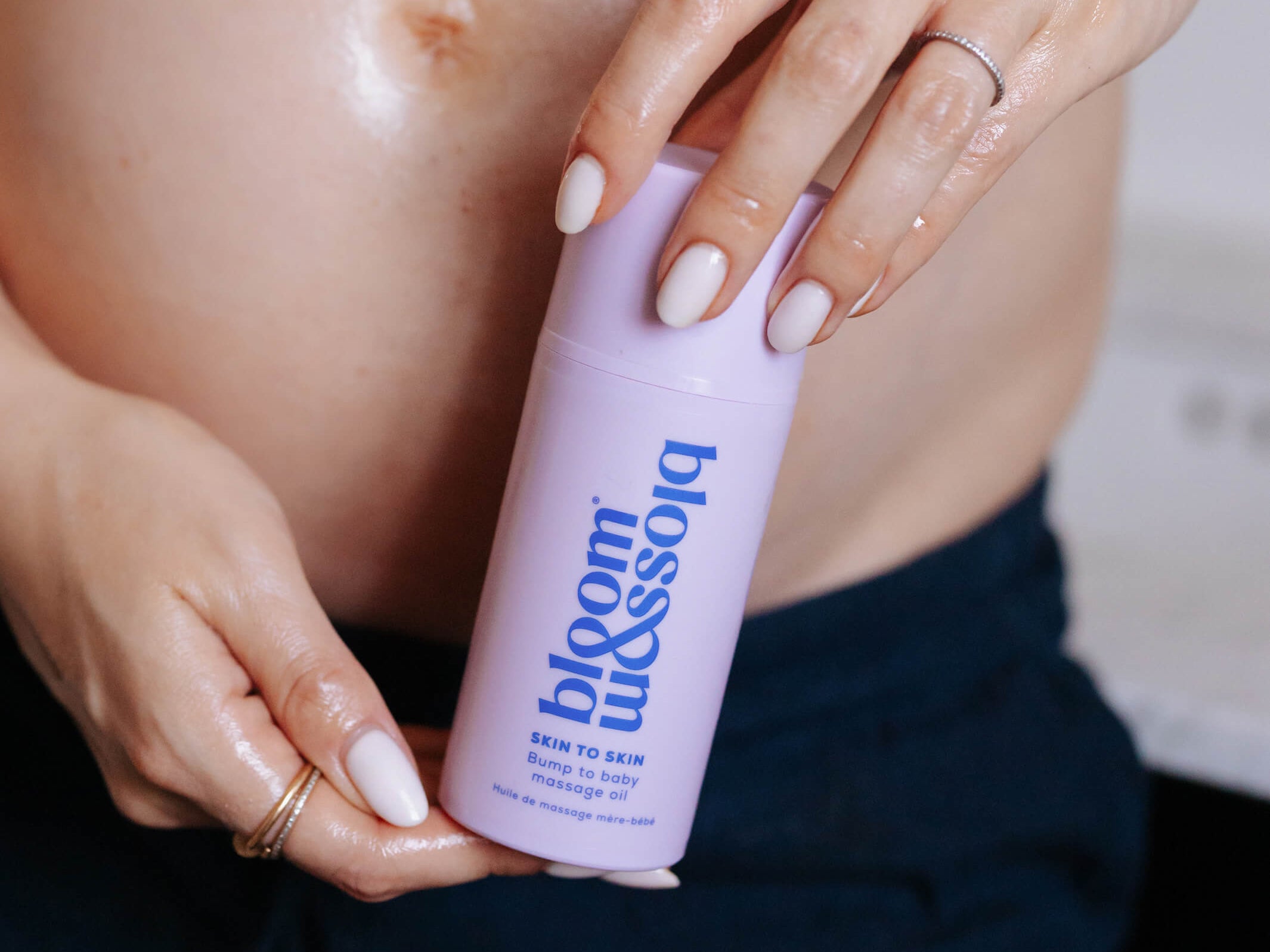 A close-up of a pregnant person holding a light purple bottle labeled 'bloom blossom' with 'skin to skin' written on it. The person is wearing a white top and dark navy pants, with their bare stomach and hands visible.