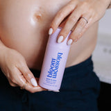 A close-up of a pregnant person holding a light purple bottle labeled 'bloom blossom' with 'skin to skin' written on it. The person is wearing a white top and dark navy pants, with their bare stomach and hands visible.