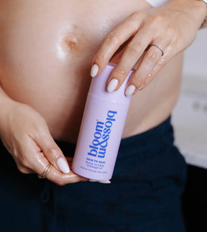 A close-up of a pregnant person holding a light purple bottle labeled 'bloom blossom' with 'skin to skin' written on it. The person is wearing a white top and dark navy pants, with their bare stomach and hands visible.