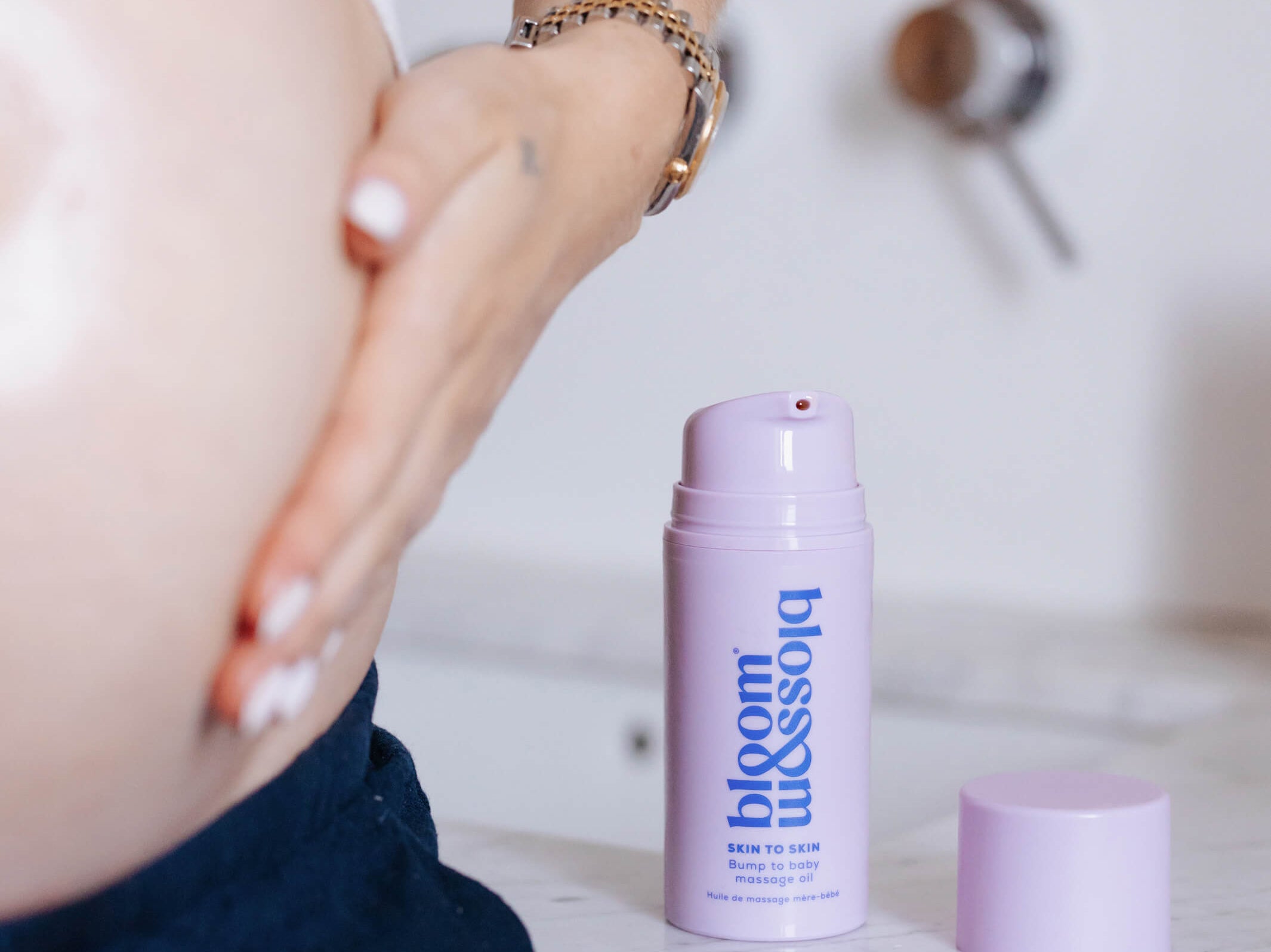 A pregnant woman's bare belly and hand are shown next to a lavender pump bottle of bloom blossom skin & hair care on a white marble countertop.