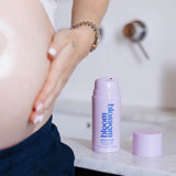A pregnant woman's bare belly and hand are shown next to a lavender pump bottle of bloom blossom skin & hair care on a white marble countertop.