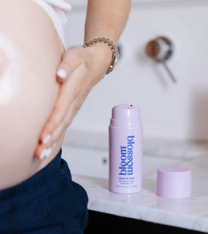 A pregnant woman's bare belly and hand are shown next to a lavender pump bottle of bloom blossom skin & hair care on a white marble countertop.