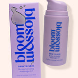 A bottle and box of bloom & blossom Skin to Skin Bump to Baby Massage Oil, shown on a warm off-white background. The packaging is lavender purple with blue text and a white droplet logo.