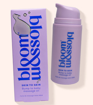 A bottle and box of bloom & blossom Skin to Skin Bump to Baby Massage Oil, shown on a warm off-white background. The packaging is lavender purple with blue text and a white droplet logo.