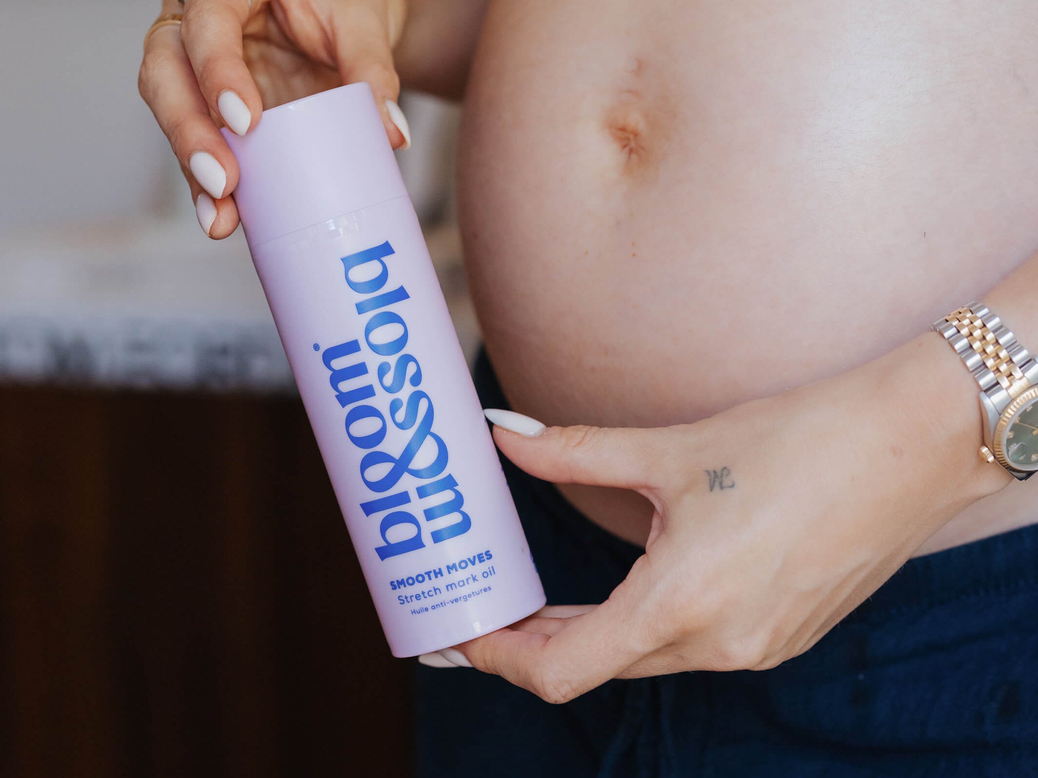 A pregnant woman holds a lavender bottle labeled 'blossom' with blue text, shown in a close-up lifestyle shot with her hands and midsection visible.