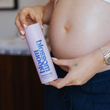 A pregnant woman holds a lavender bottle labeled 'blossom' with blue text, shown in a close-up lifestyle shot with her hands and midsection visible.