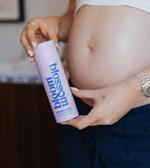 A pregnant woman holds a lavender bottle labeled 'blossom' with blue text, shown in a close-up lifestyle shot with her hands and midsection visible.