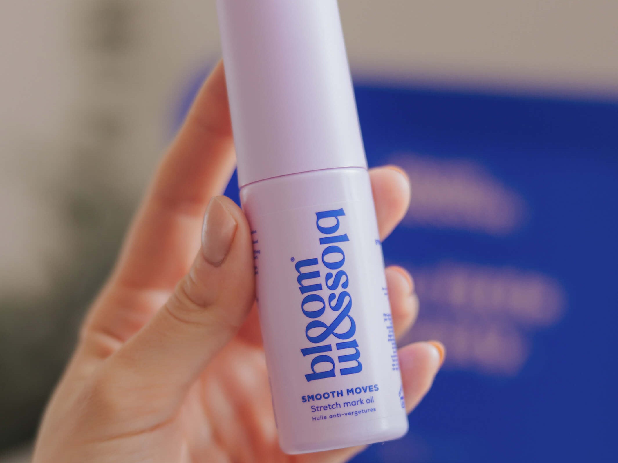 A hand holding a bottle of blossom beauty Smooth Moves Smoothing Mask Oil against a soft-focus background with a prominent blue box.