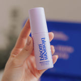 A hand holding a bottle of blossom beauty Smooth Moves Smoothing Mask Oil against a soft-focus background with a prominent blue box.