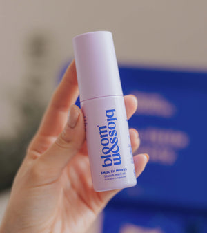 A hand holding a bottle of blossom beauty Smooth Moves Smoothing Mask Oil against a soft-focus background with a prominent blue box.