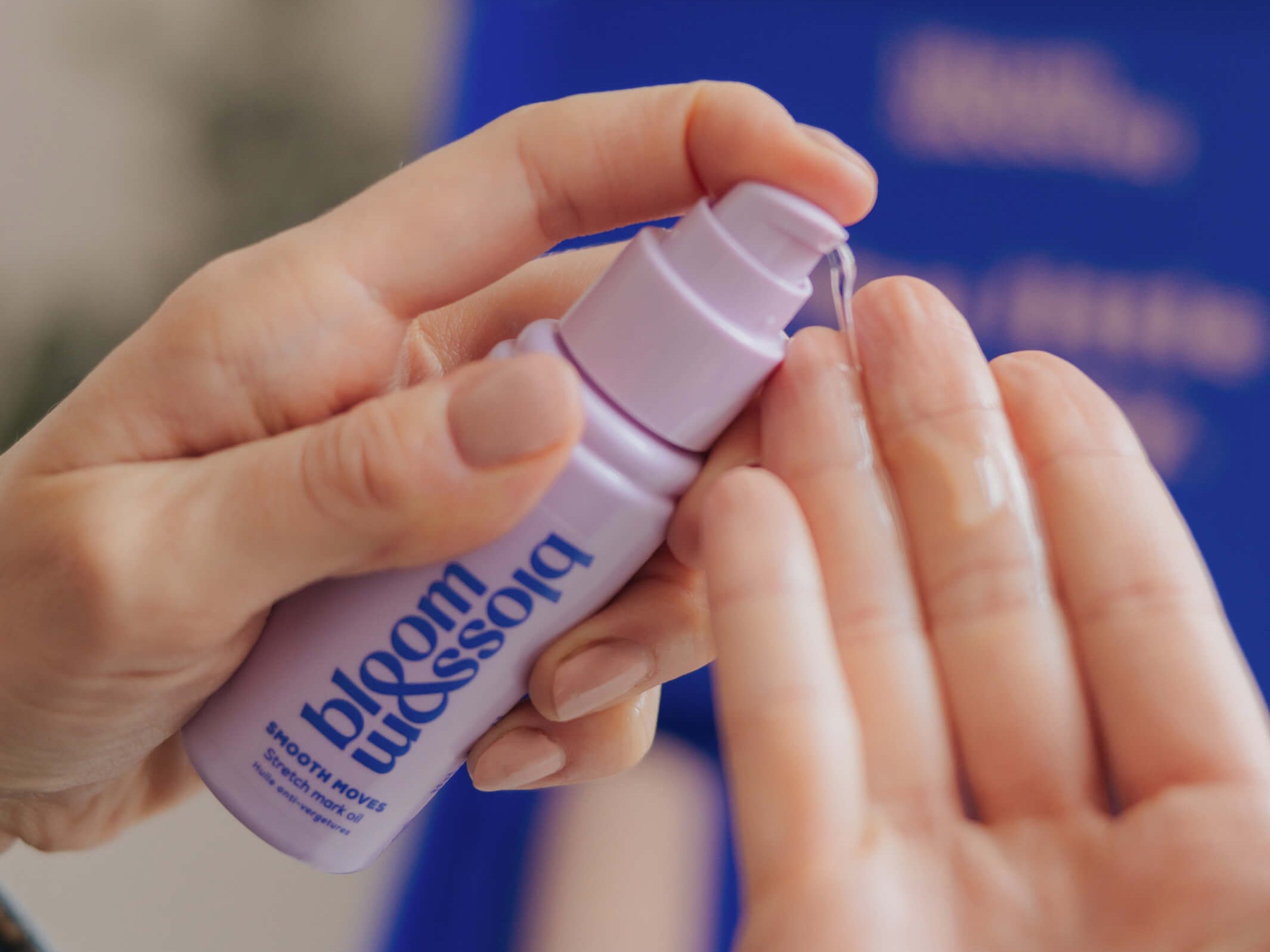 A close-up of hands applying a clear liquid from a small lavender pump bottle labeled bloom blossoms. The background is softly blurred with a blue box and neutral indoor tones.