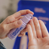 A close-up of hands applying a clear liquid from a small lavender pump bottle labeled bloom blossoms. The background is softly blurred with a blue box and neutral indoor tones.