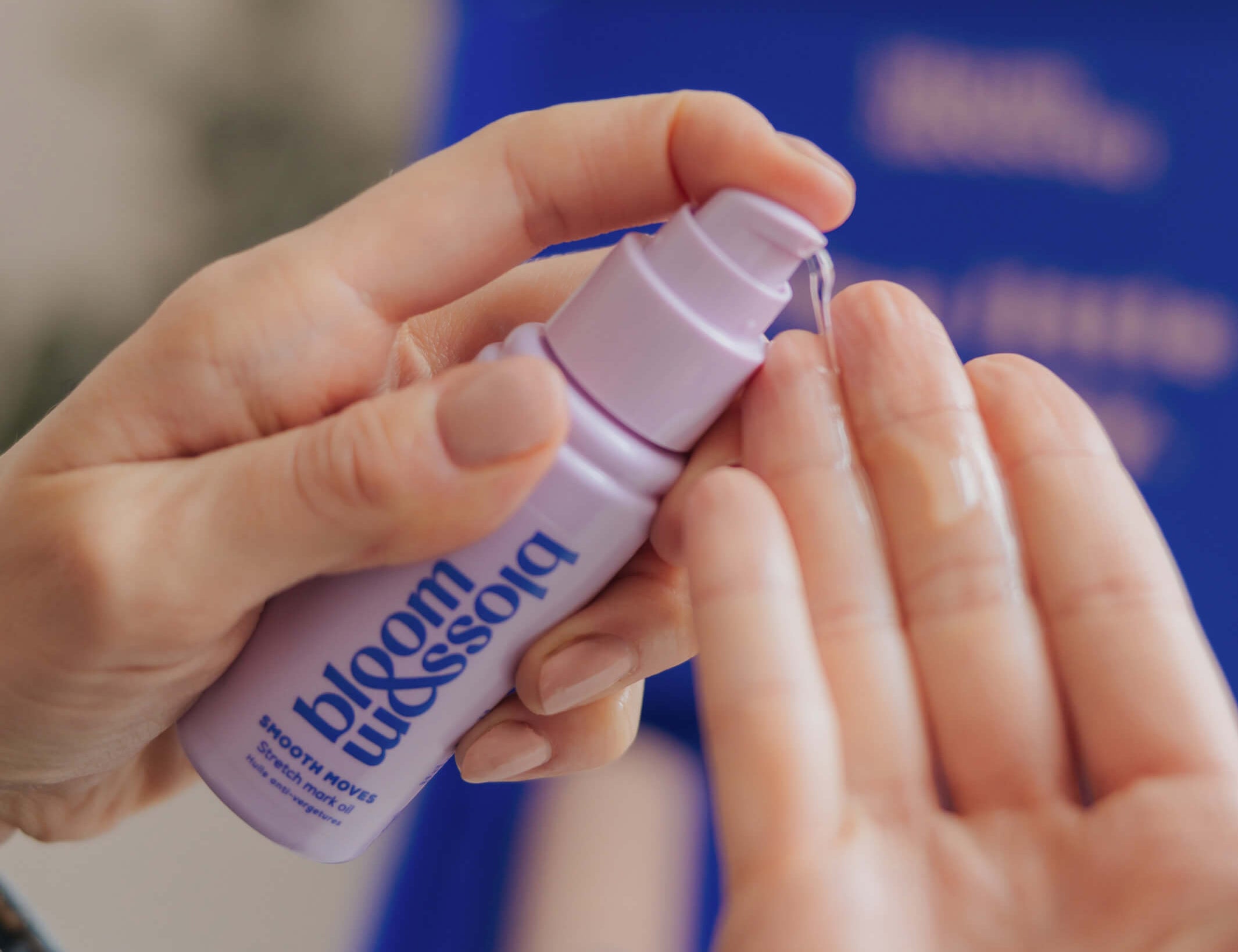 A close-up of hands applying a clear liquid from a small lavender pump bottle labeled bloom blossoms. The background is softly blurred with a blue box and neutral indoor tones.