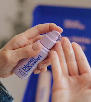 A close-up of hands applying a clear liquid from a small lavender pump bottle labeled bloom blossoms. The background is softly blurred with a blue box and neutral indoor tones.