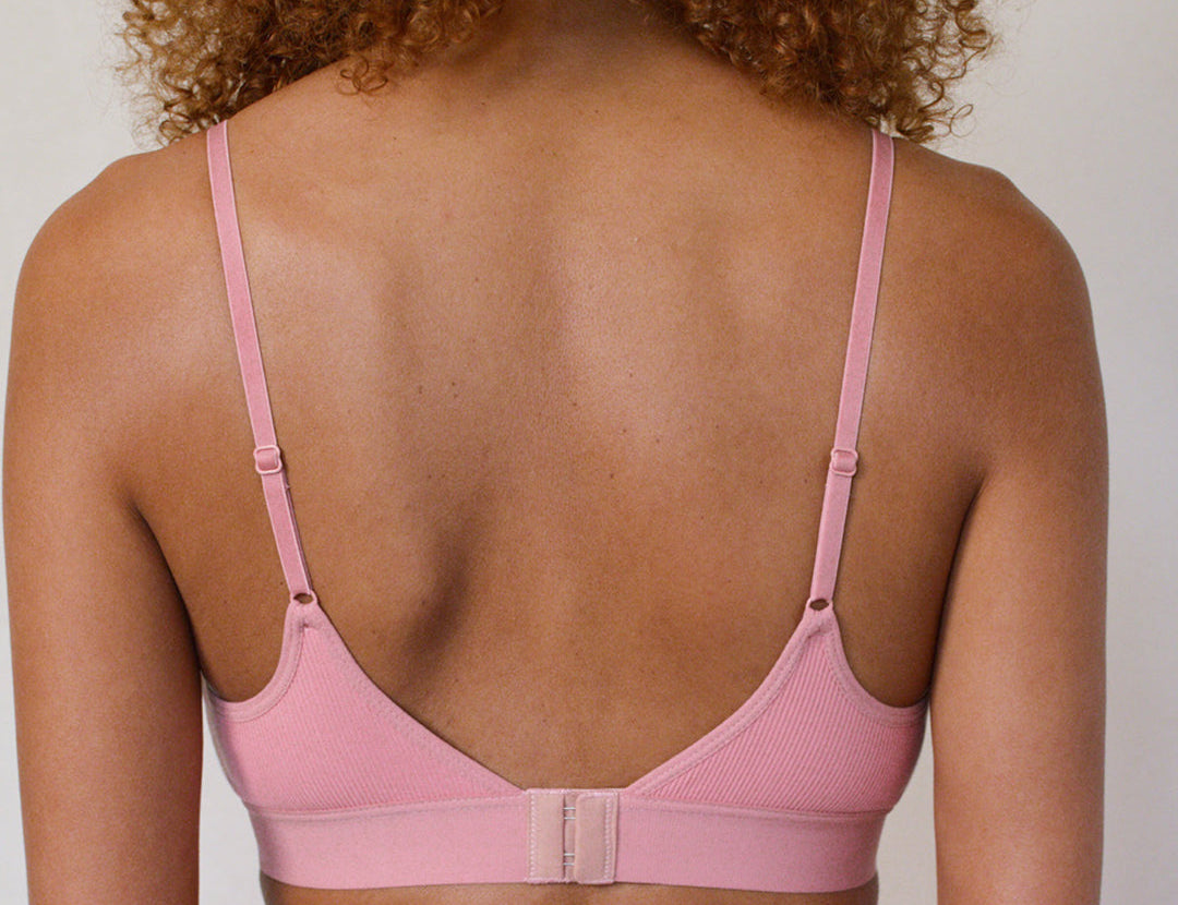 Back view of a woman wearing a light pink ribbed underwire bra with thin adjustable shoulder straps and a center back strap connector, shown against a plain light gray background.