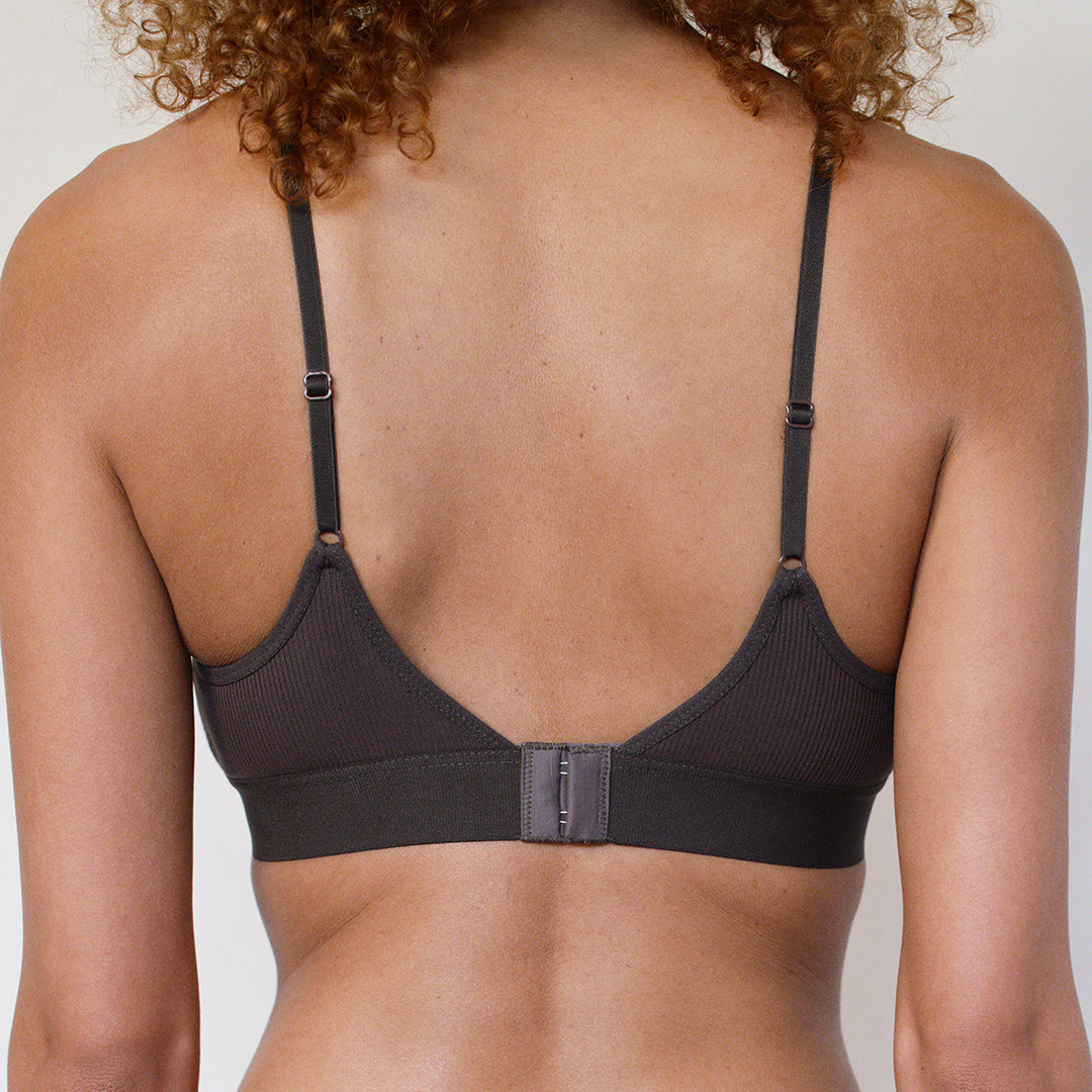 Back view of a woman wearing a dark charcoal ribbed underwire bra with adjustable shoulder straps and a center hook-and-loop closure.