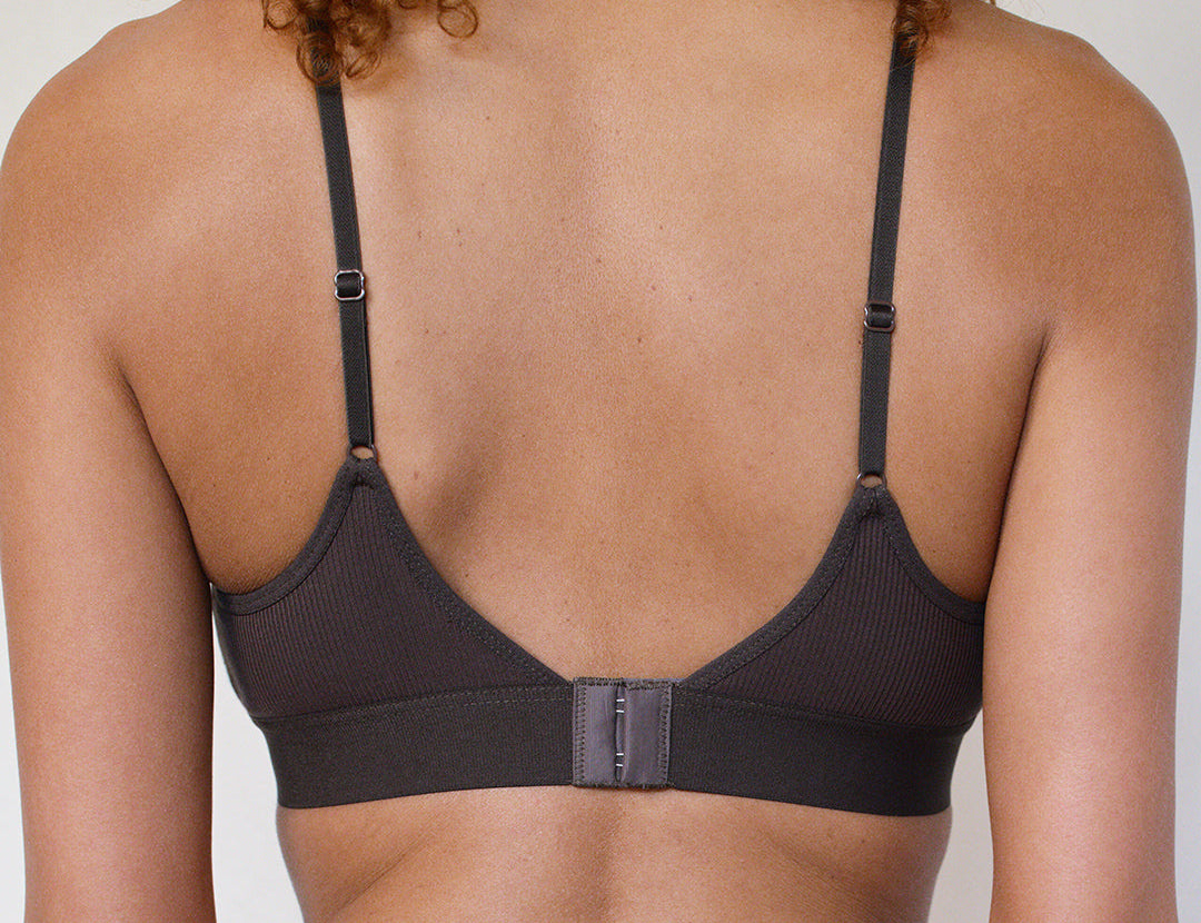 Back view of a woman wearing a dark charcoal ribbed underwire bra with adjustable shoulder straps and a center hook-and-loop closure.