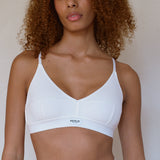A close-up of a model wearing a white ribbed wireless bra with underwire support and a small 'RENUE' brand label on the front band, set against a plain light beige wall.
