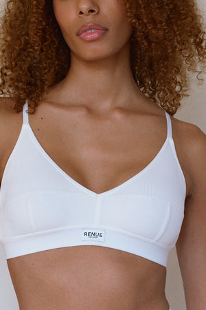 A close-up of a model wearing a white ribbed wireless bra with underwire support and a small 'RENUE' brand label on the front band, set against a plain light beige wall.