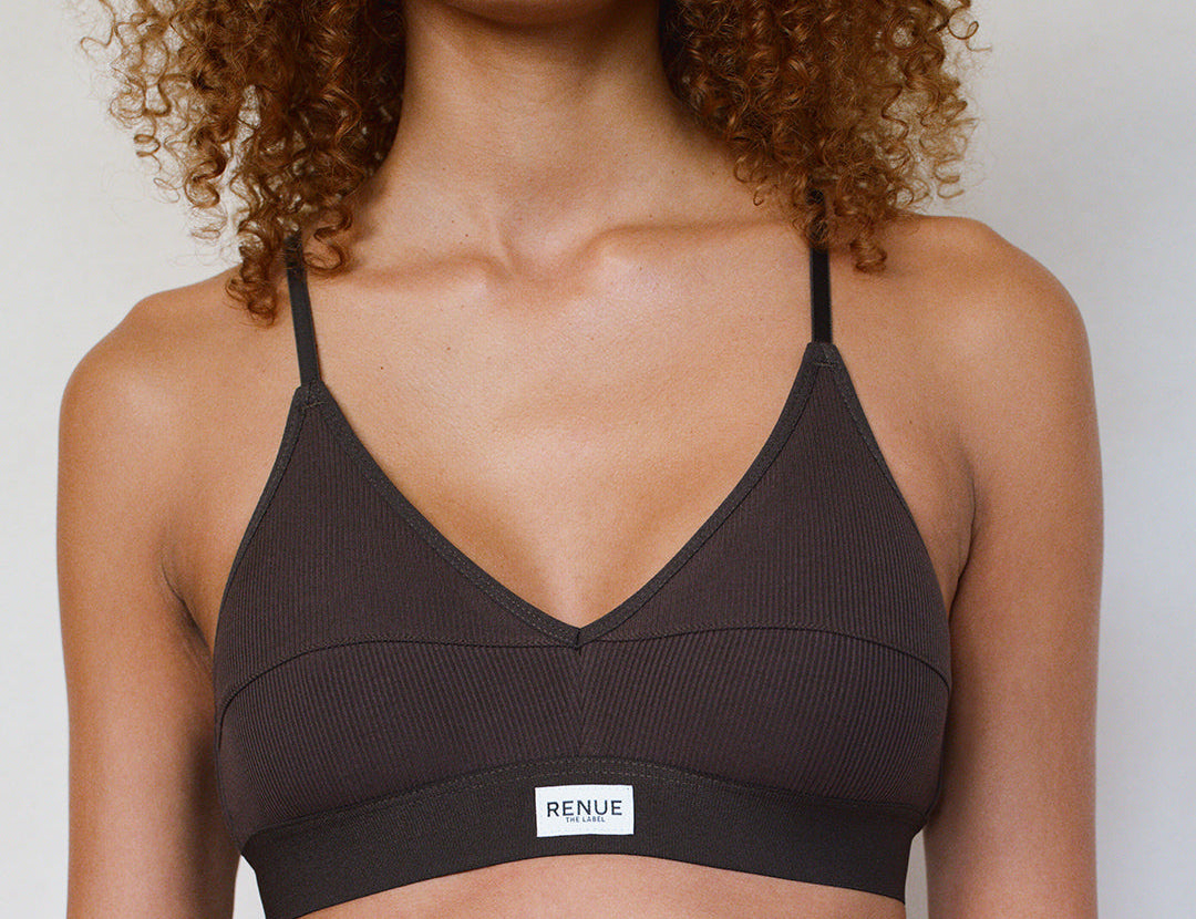 A close-up of a model wearing a dark brown ribbed V-neck bralette with thin straps and a white brand label that reads 'RENUE'.