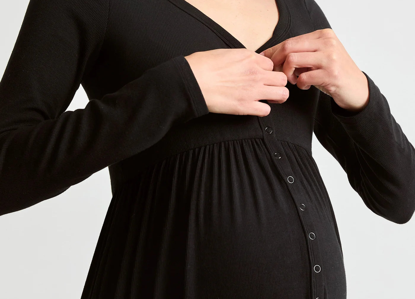 A pregnant woman wearing a black long-sleeve maternity dress with a buttoned front panel, shown from the chest down against a light gray studio background.