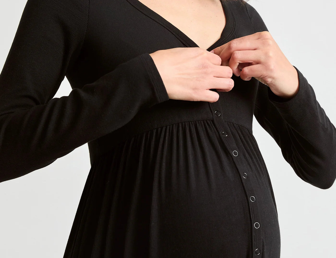 A pregnant woman wearing a black long-sleeve maternity dress with a buttoned front panel, shown from the chest down against a light gray studio background.
