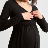 A pregnant woman wearing a black long-sleeve maternity dress with a buttoned front panel, shown from the chest down against a light gray studio background.