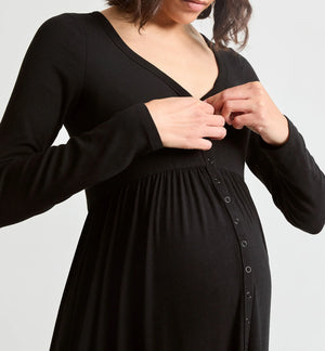 A pregnant woman wearing a black long-sleeve maternity dress with a buttoned front panel, shown from the chest down against a light gray studio background.