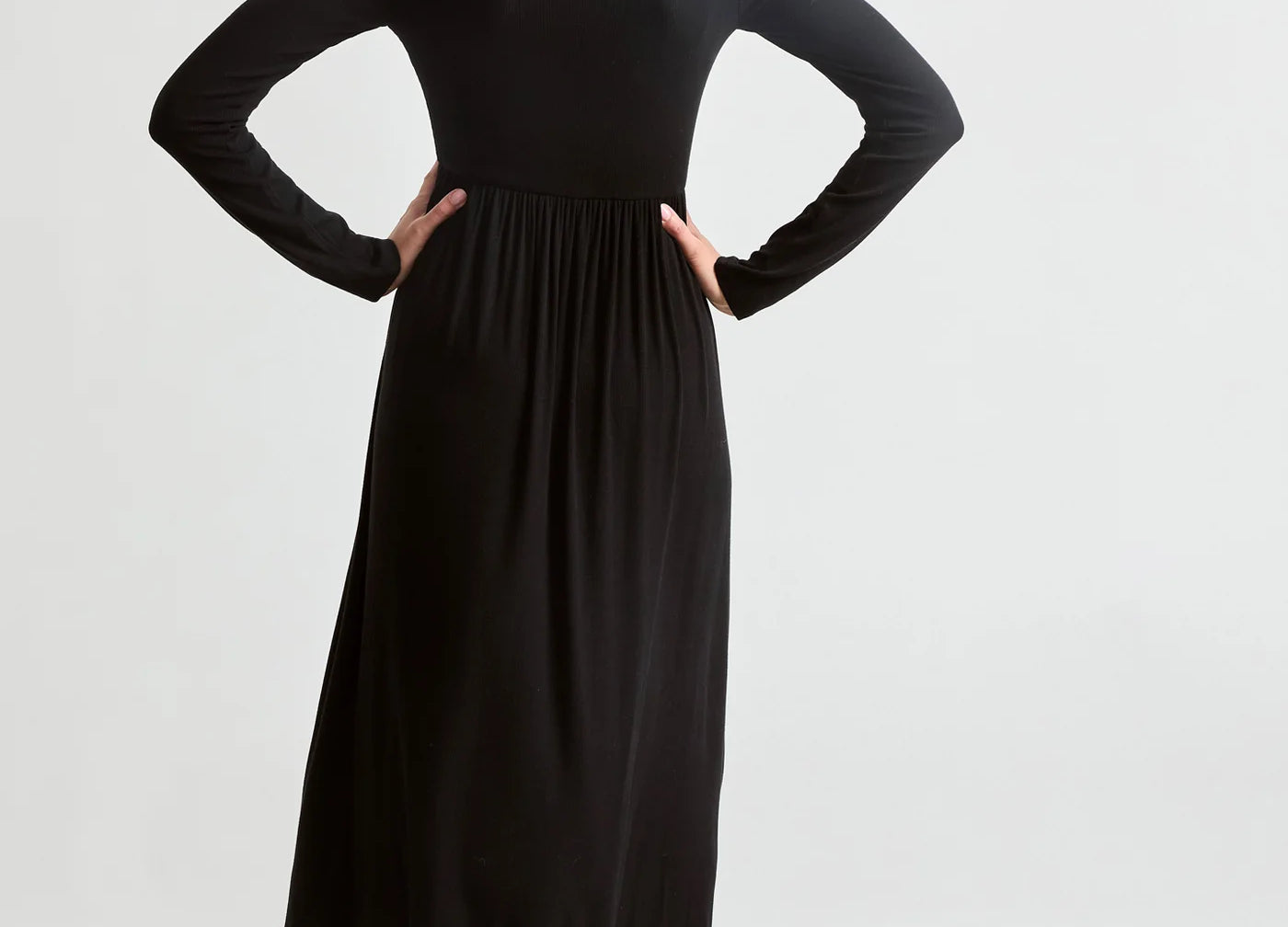 A woman shown from the back wearing a black long-sleeve midi dress with a gathered waist, paired with black ankle boots, against a plain light gray studio background.