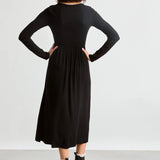 A woman shown from the back wearing a black long-sleeve midi dress with a gathered waist, paired with black ankle boots, against a plain light gray studio background.