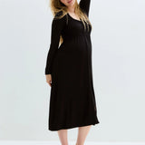 A pregnant woman modeling a black long-sleeve maternity maxi dress, standing barefoot against a clean white studio background.