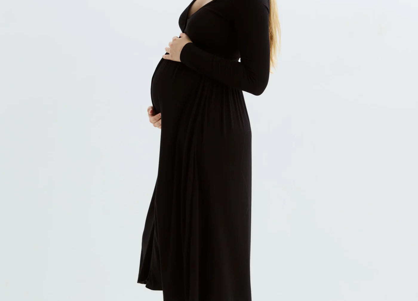A pregnant woman with blonde hair stands in profile wearing a black long-sleeve maternity dress, standing barefoot against a clean white studio background.