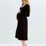 A pregnant woman with blonde hair stands in profile wearing a black long-sleeve maternity dress, standing barefoot against a clean white studio background.