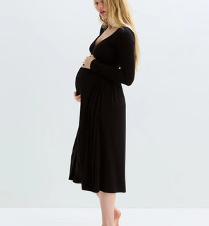 A pregnant woman with blonde hair stands in profile wearing a black long-sleeve maternity dress, standing barefoot against a clean white studio background.