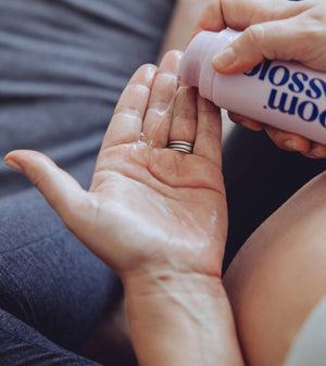 A close-up of a person pouring a clear liquid from a pink bottle labeled 'blossom wood' onto the back of their hand.