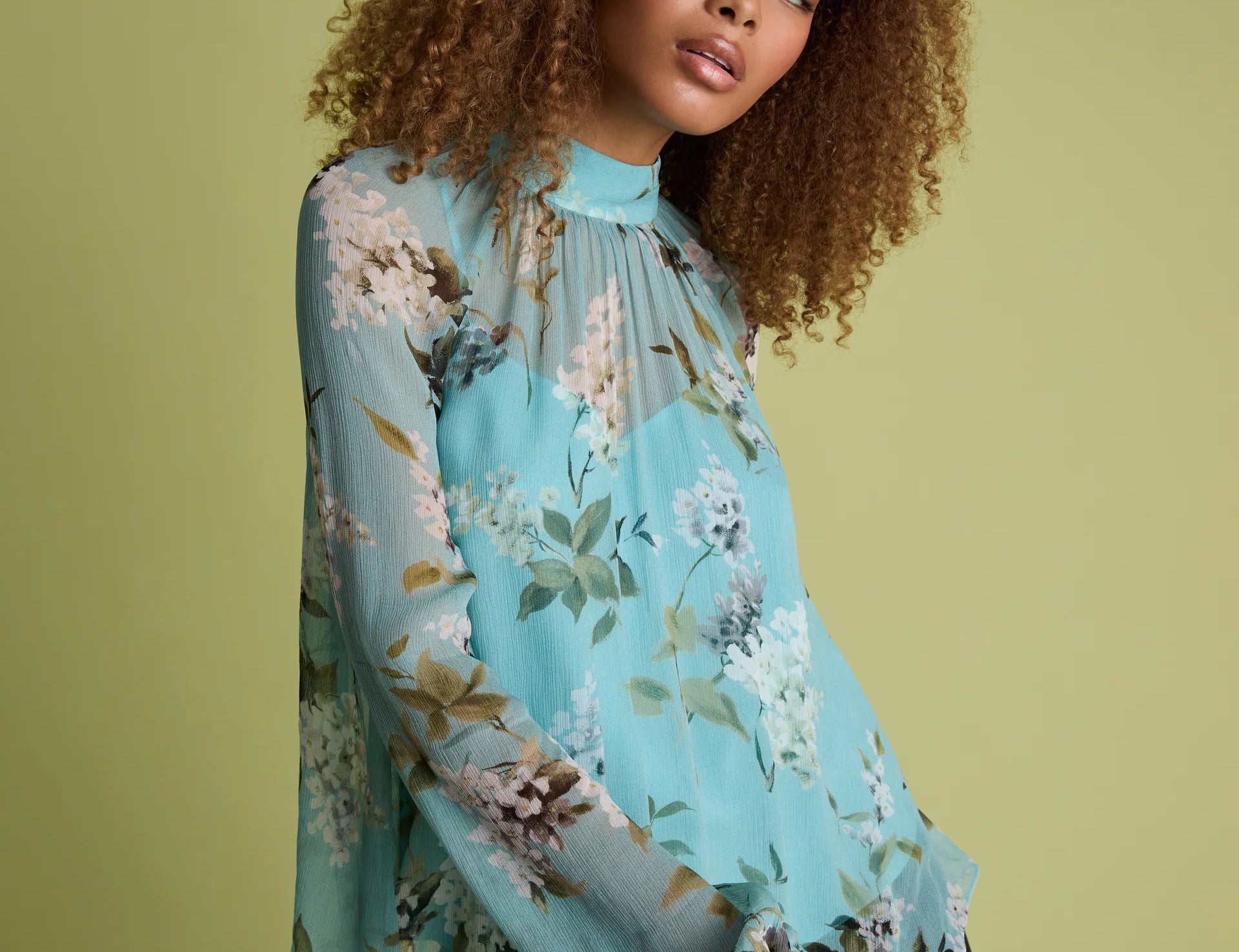 A woman with curly brown hair wearing a light blue floral long-sleeve blouse and black pants, posing against a solid olive green background.