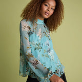 A woman with curly brown hair wearing a light blue floral long-sleeve blouse and black pants, posing against a solid olive green background.