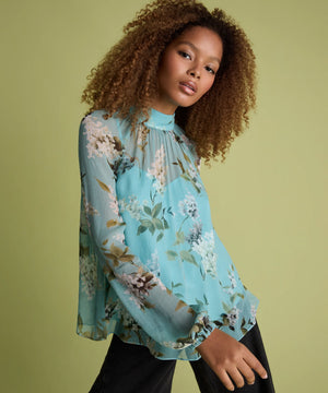 A woman with curly brown hair wearing a light blue floral long-sleeve blouse and black pants, posing against a solid olive green background.