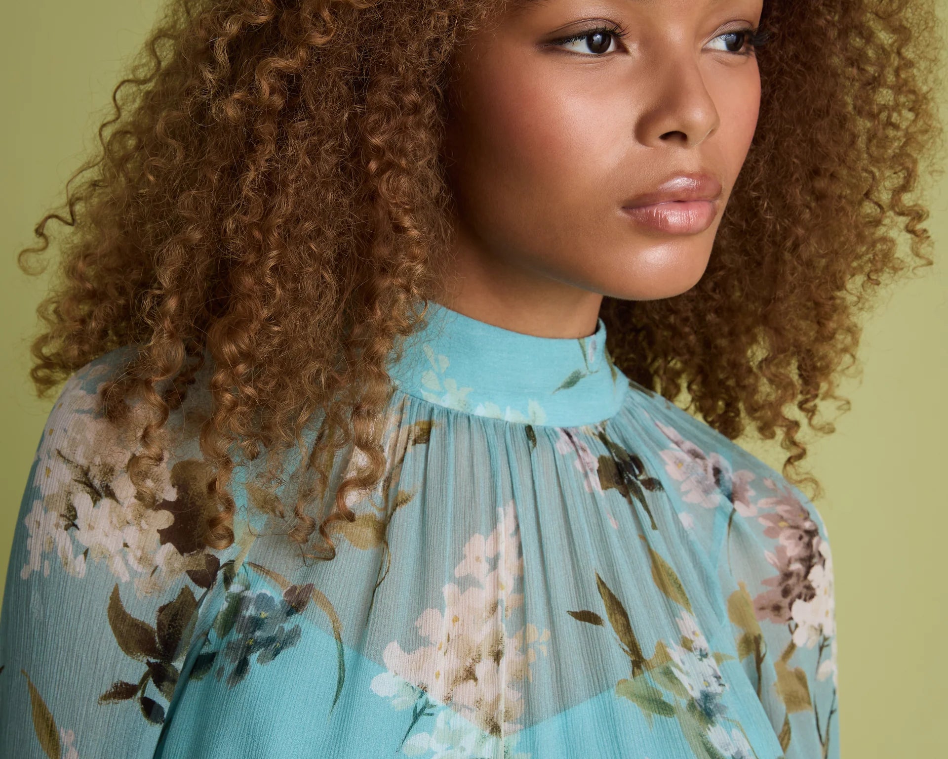 A close-up portrait of a woman with voluminous curly brown hair wearing a light blue high-neck blouse with a floral print, set against a solid olive green background.