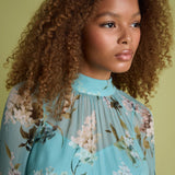 A close-up portrait of a woman with voluminous curly brown hair wearing a light blue high-neck blouse with a floral print, set against a solid olive green background.