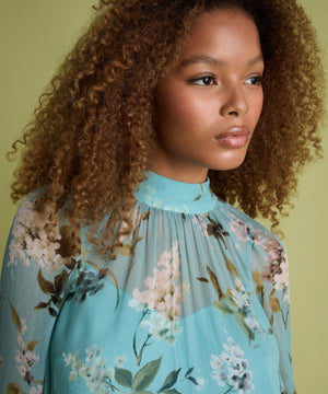 A close-up portrait of a woman with voluminous curly brown hair wearing a light blue high-neck blouse with a floral print, set against a solid olive green background.