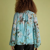A person shown from the back wearing a light blue floral long-sleeve blouse with a tie neckline and dark jeans, standing against a solid olive-green background.