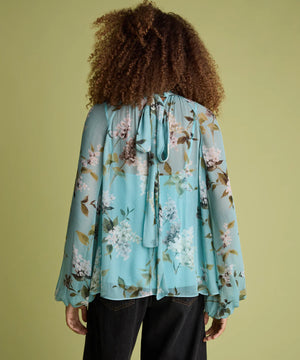 A person shown from the back wearing a light blue floral long-sleeve blouse with a tie neckline and dark jeans, standing against a solid olive-green background.