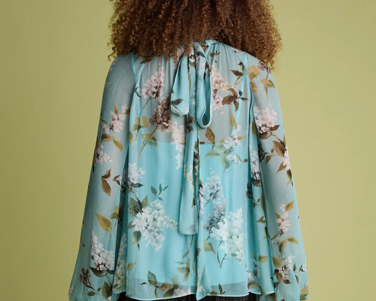 A person shown from the back wearing a light blue floral long-sleeve blouse with a tie neckline and dark jeans, standing against a solid olive-green background.