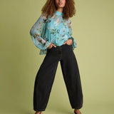 A model with curly hair stands against a solid sage green studio background, wearing a light blue floral long-sleeve blouse with ruffled cuffs, black wide-leg cropped jeans, and black pointed-toe heels.