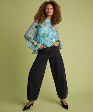 A model with curly hair stands against a solid sage green studio background, wearing a light blue floral long-sleeve blouse with ruffled cuffs, black wide-leg cropped jeans, and black pointed-toe heels.