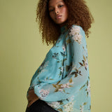 A woman with curly brown hair models a light blue floral long-sleeve blouse with black jeans against a solid light olive green background.